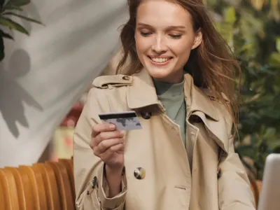 Femeie zâmbitoare ținând în mână un card de credit și zâmbind, în timp ce lucrează la laptop, într-o cafenea cu terasă - Foto: Magnific.com/garetsvisual