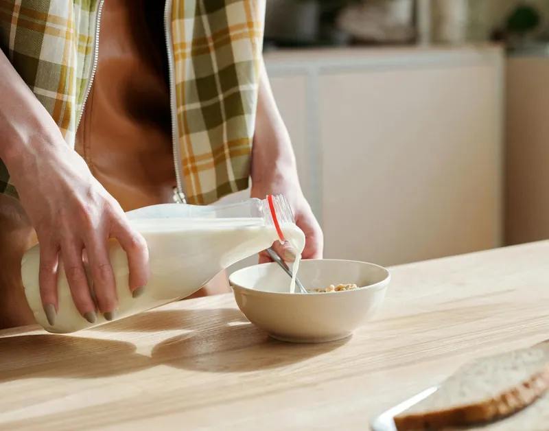 Femeie turn&acirc;nd lapte dintr-o sticlă, &icirc;ntr-un bol, &icirc;n bucătărie - Foto: Pexels/Mikael Blomkvist