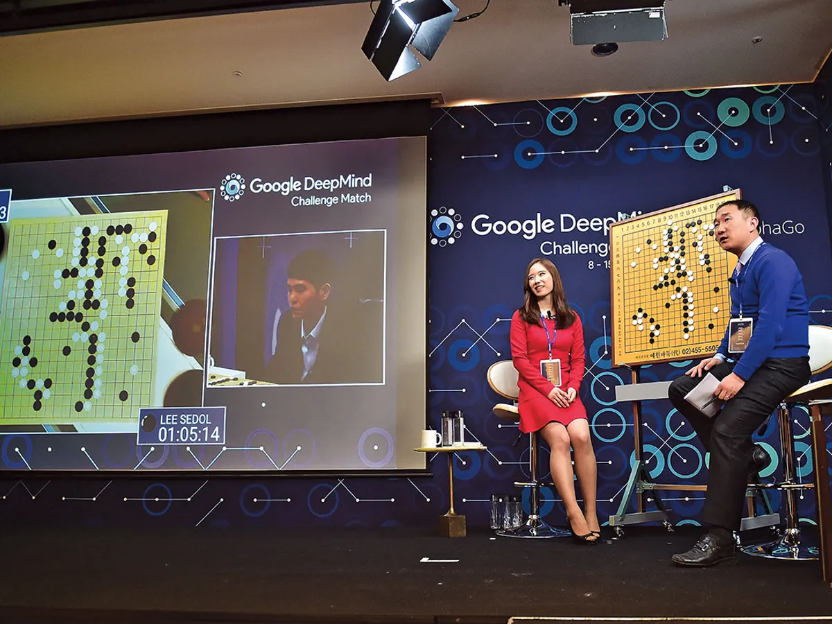 Comentatorii explică, în timp ce, pe ecran, marele maestru sud-coreean la Go Lee Se-Dol provoacă supercomputerul AlphaGo în 2016; Ke Jie, în vârstă de 19 ani, se confruntă cu AlphaGo în 2017