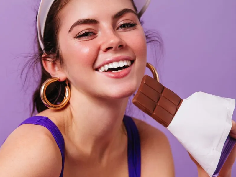 Tânără fată zâmbitoare muşcând dintr--o ciocolată - Foto: Freepik/lookstudio