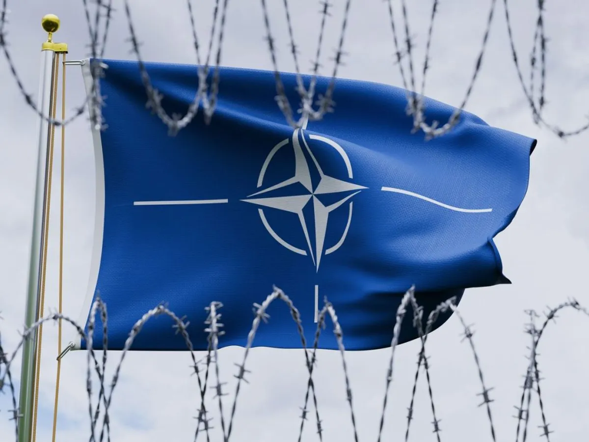 Drapelul NATO flutură mândru, pe un catarg, privit printr-o gard de sârmă ghimpată, pe fundalul unui cer înnorat - Foto: Profimedia Images