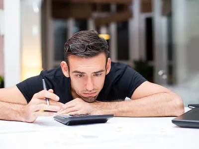 Bărbat stând cu bărbia pe birou şi făcând calcule pe laptop - Foto: Magnific.com/katemangostar
