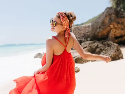 Femeie zâmbitoare, pe plajă, în rochie vaporoasă, roşie - Foto: Freepik/lookstudio