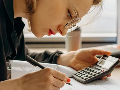 Femeie scriind într-o agendă, în timp ce ţine în mână un calculator - Foto: Pexels/Mikhail Nilov