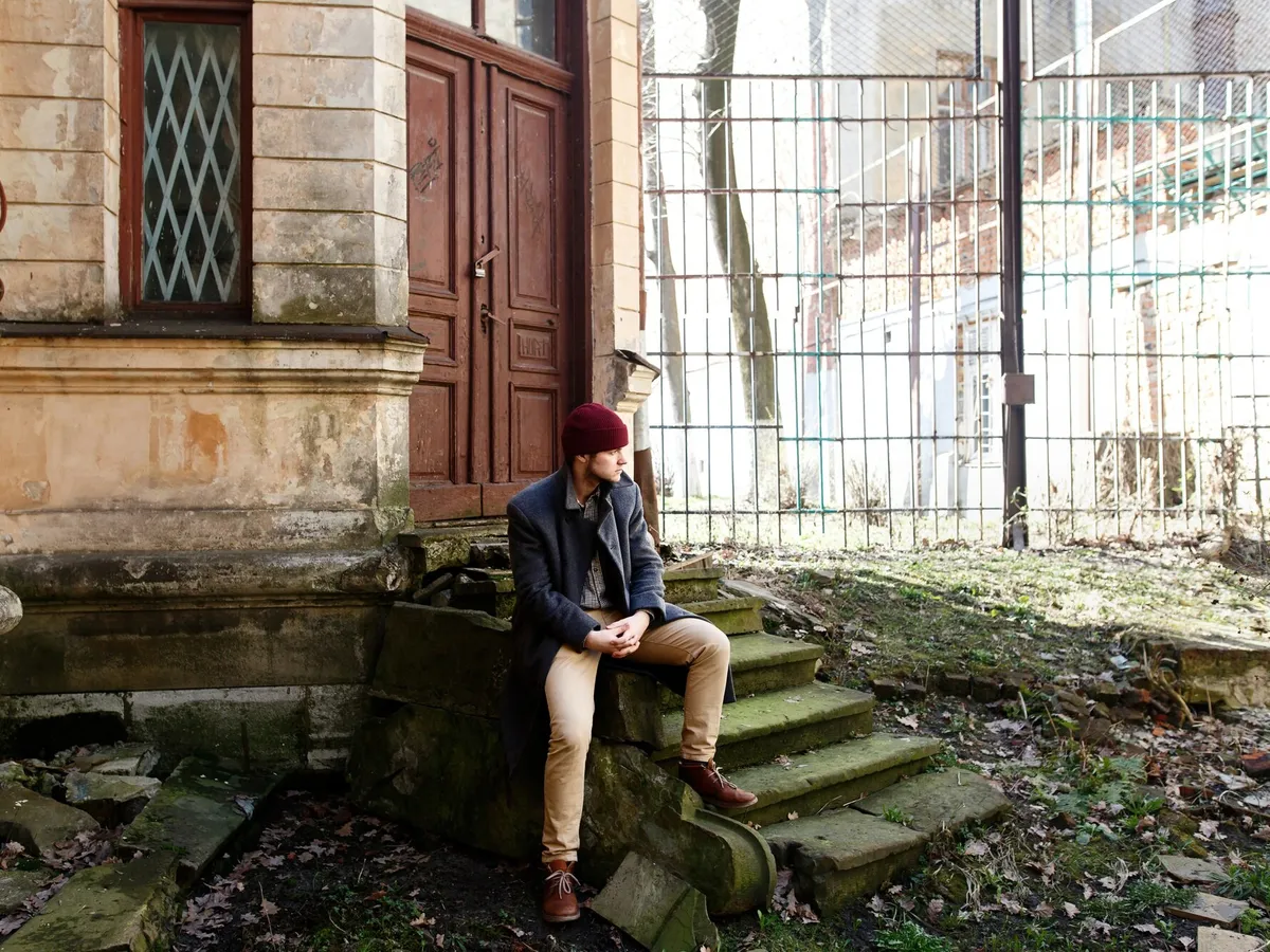 Bărbat cu pălărie roșie și haină gri stă pe scările din fața unei case vechi - Foto: Freepik (imagine cu rol ilustrativ)