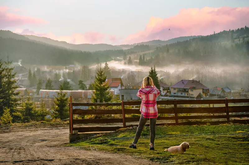 Vedere din spate a unei femei și a unui câine care privesc satul încețoșat la apus - Foto: Freepik (rol ilustrativ)