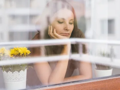 Femeie frumoasă care se uită tristă pe fereastră din casă - Foto: Freepik