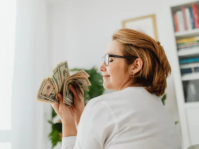 Femeie încrezătoare cu ochelari, ținând bancnote în dolari americani - Foto: Pexels (Imagine cu rol ilustrativ)