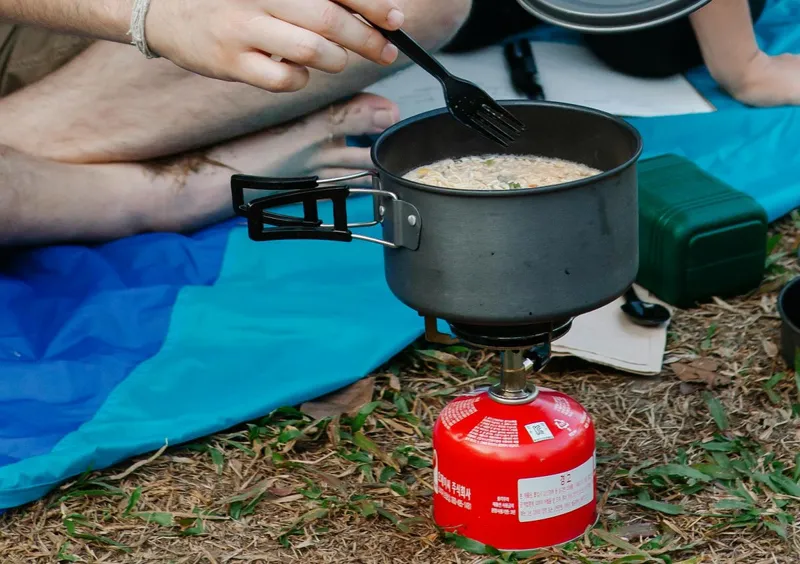 Cuplu la iarbă verde &icirc;ncălzindu-şi dejunul la o butelie autonomă de gaz - Foto: Pexels/Vanessa Garcia