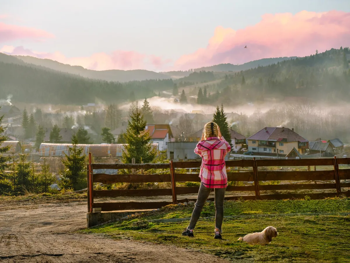 Vedere din spate a unei femei și a unui câine care privesc satul încețoșat la apus - Foto: Freepik (rol ilustrativ)