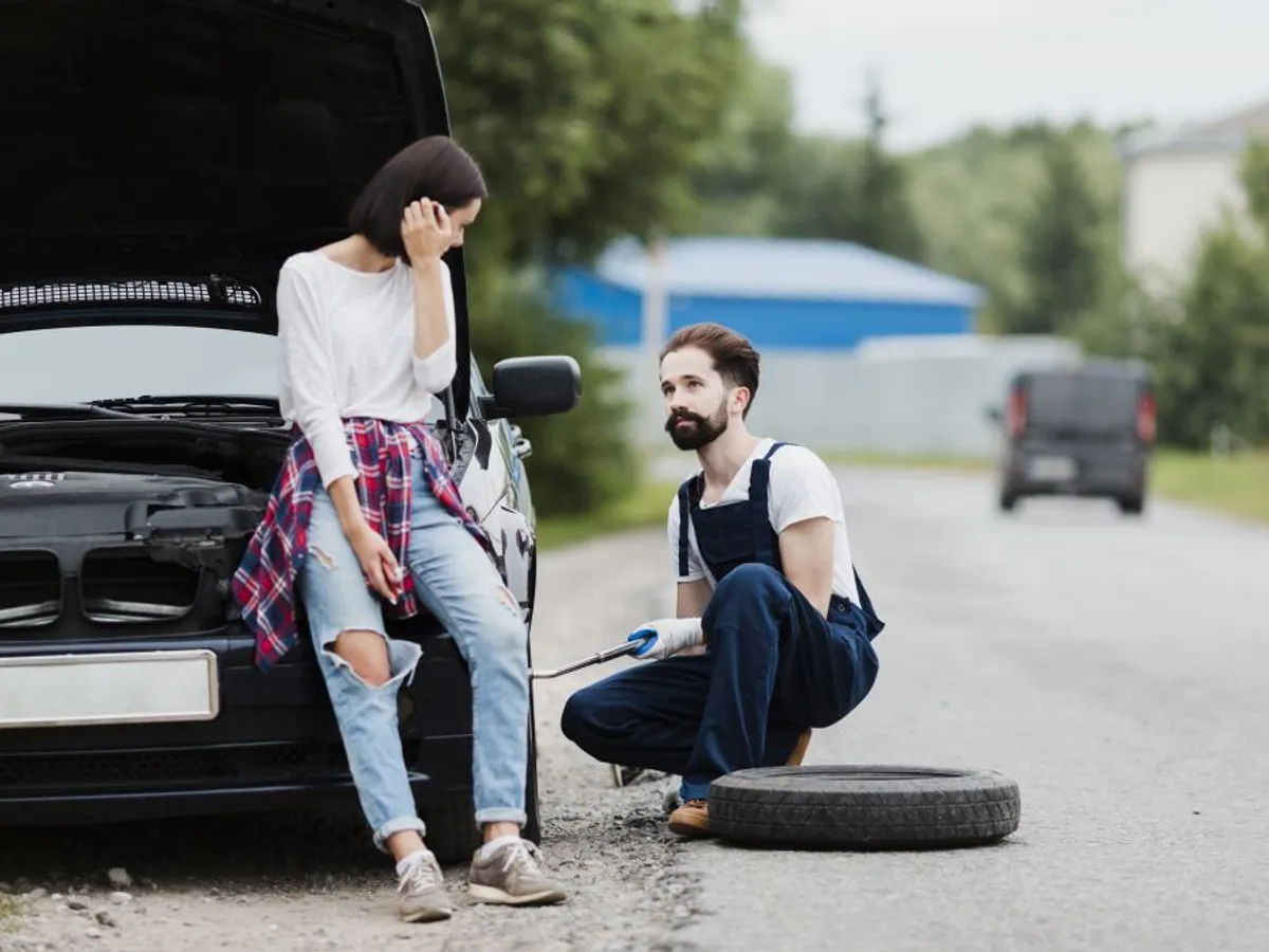 O femeie stă sprijinită de botul maşinii, după un accident, în timp ce un bărbat îi schimbă anvelopa - Foto: Freepik/CC0
