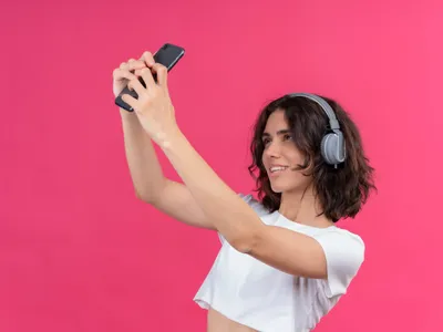 Tânără frumoasă zâmbitoare purtând căști și ținând telefonul mobil făcându-și selfie-uri - Foto: Magnific.com (Imagine cu rol ilustrativ)