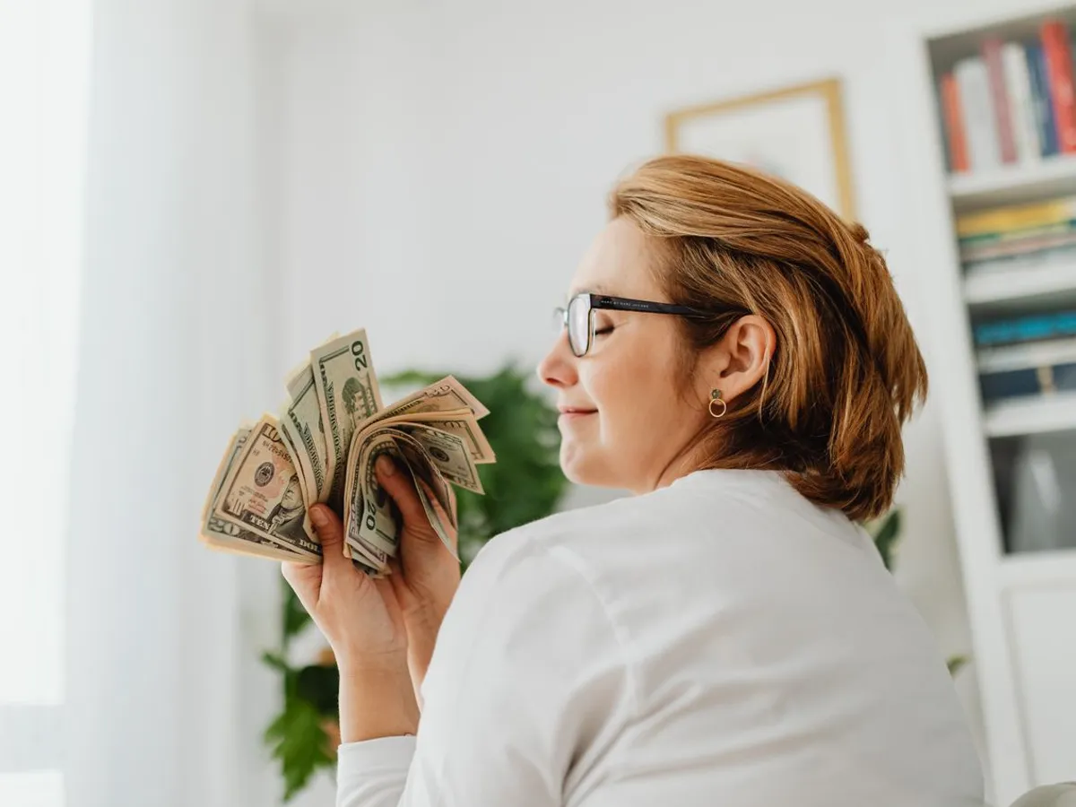 Femeie încrezătoare cu ochelari, ținând bancnote în dolari americani - Foto: Pexels (Imagine cu rol ilustrativ)