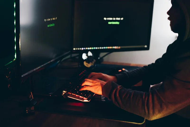Silueta unui bărbat care scrie  la tastatură, &icirc;n faţa a două monitoare - Foto: Pexels/Anete Lusina