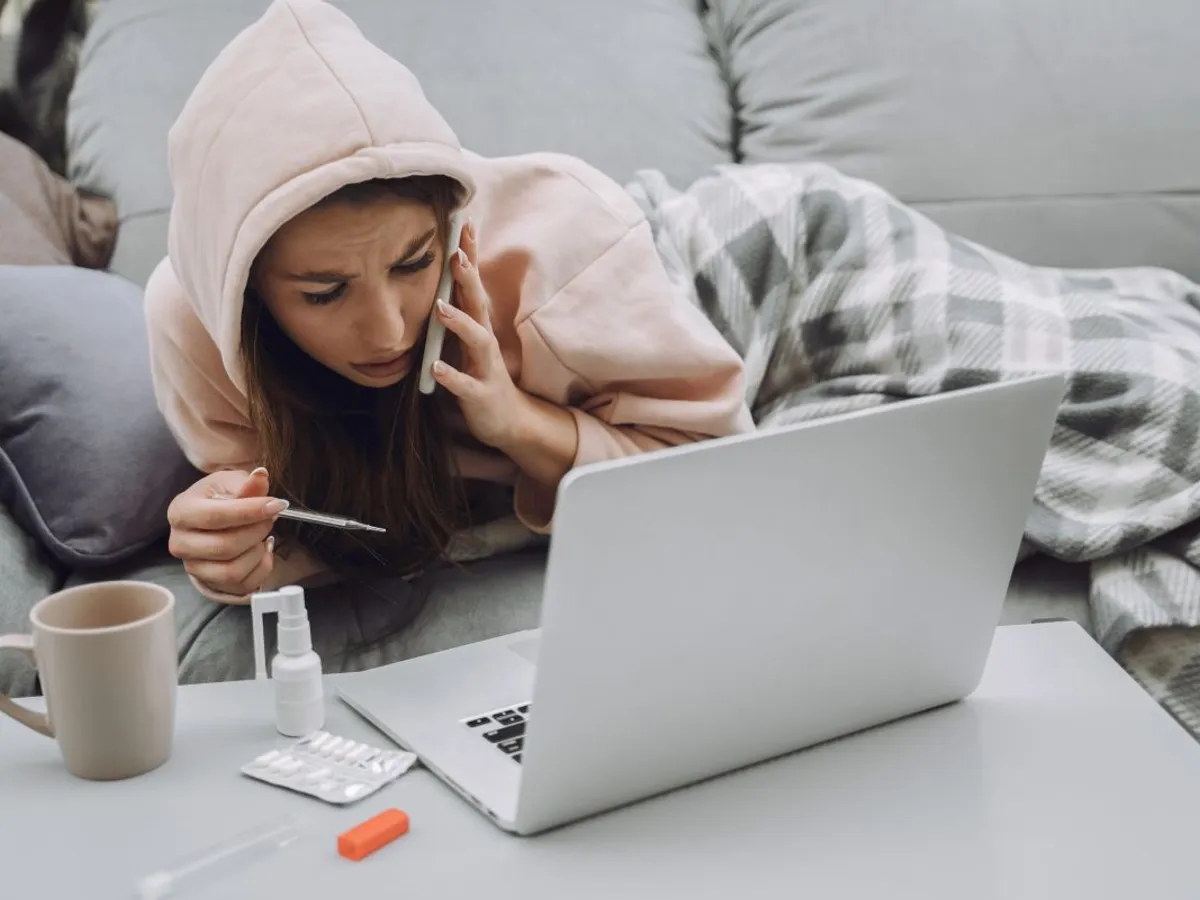 Femeie răcită, în pat, vorbind la telefon, de sub pătură, cu laptopul în faţă - Foto: Magnific.com/prostooleh