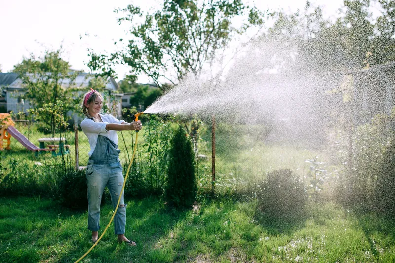 O t&acirc;nără femeie &icirc;ntr-o cămașă albă și o salopetă din denim udă gazonul - Foto: Magnific.com (Imagine cu rol ilustrativ)