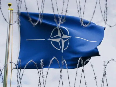 Drapelul NATO flutură mândru, pe un catarg, privit printr-o gard de sârmă ghimpată, pe fundalul unui cer înnorat - Foto: Profimedia Images