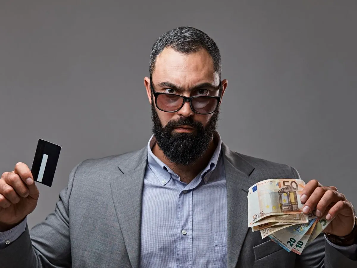 Bărbat bărbos ţinând în mâna dreaptă un card bancar şi în stânga bancnote euro - Foto: Magnific.com/fxquadro