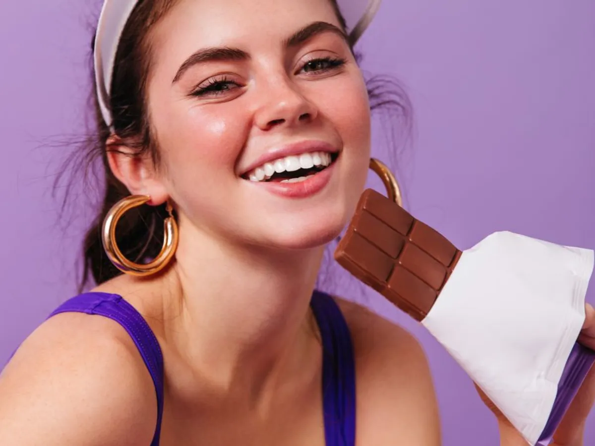 Tânără fată zâmbitoare muşcând dintr--o ciocolată - Foto: Freepik/lookstudio