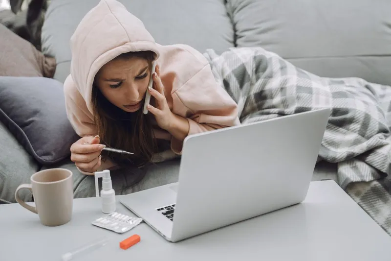 Femeie răcită, &icirc;n pat, vorbind la telefon, de sub pătură, cu laptopul &icirc;n faţă - Foto: Magnific.com/prostooleh