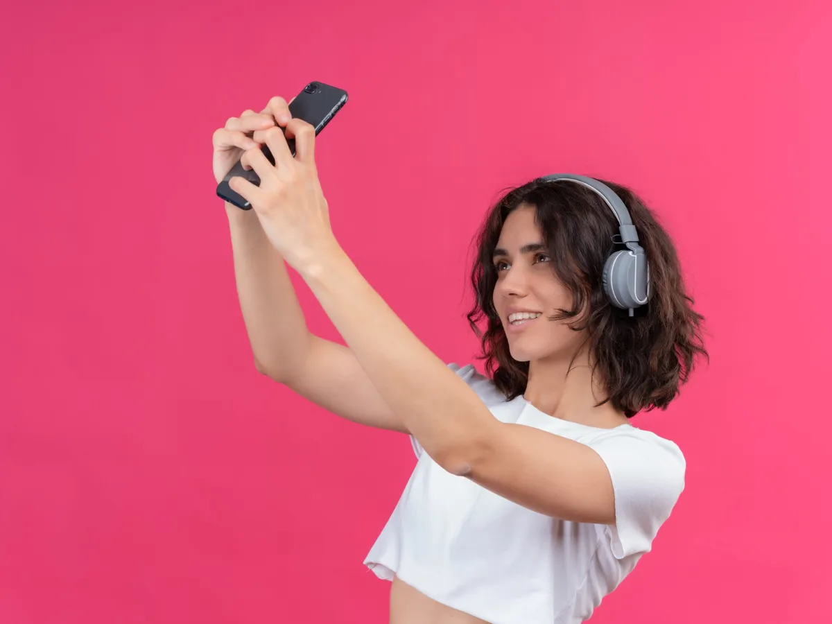 Tânără frumoasă zâmbitoare purtând căști și ținând telefonul mobil făcându-și selfie-uri - Foto: Magnific.com (Imagine cu rol ilustrativ)