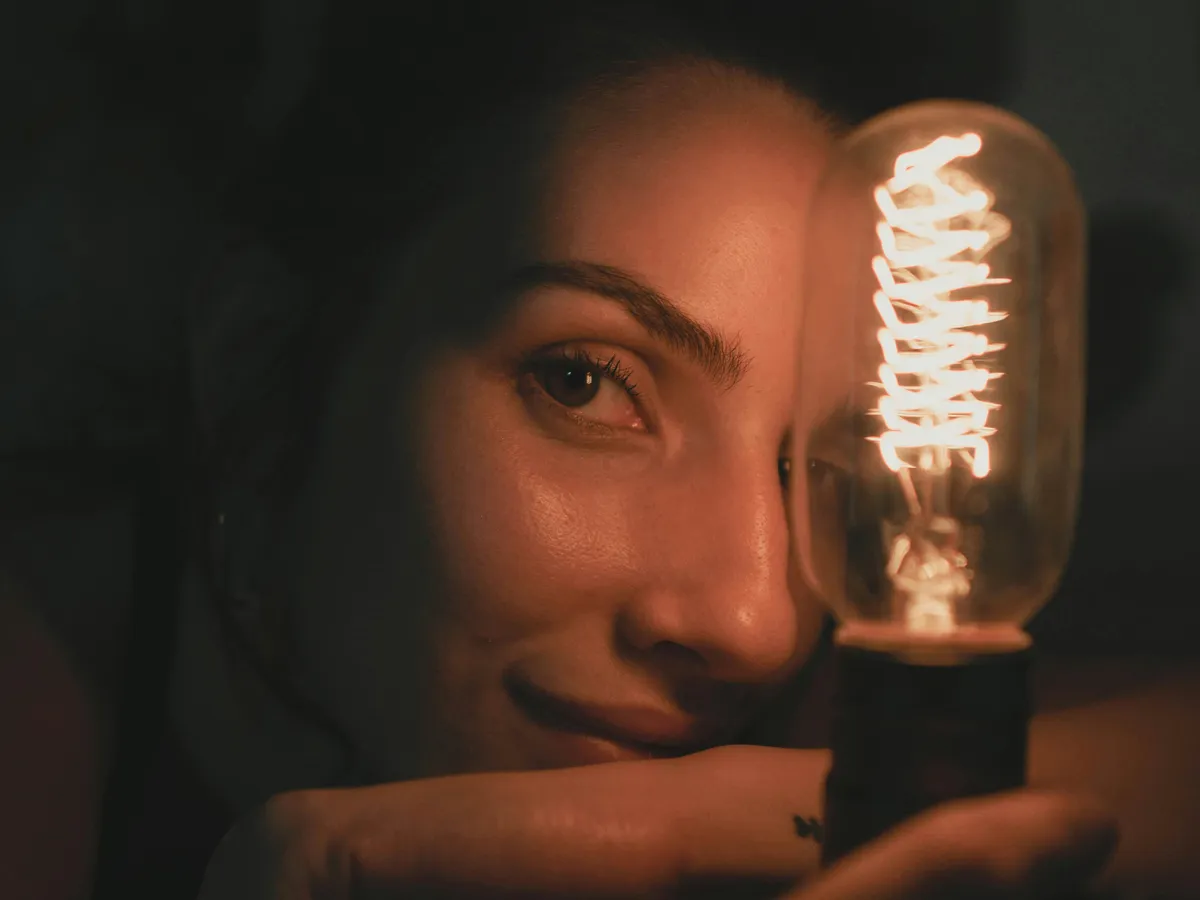 Tânără femeie ţinând în mână un bec cu filament - Foto: Pexels/Caique Araujo