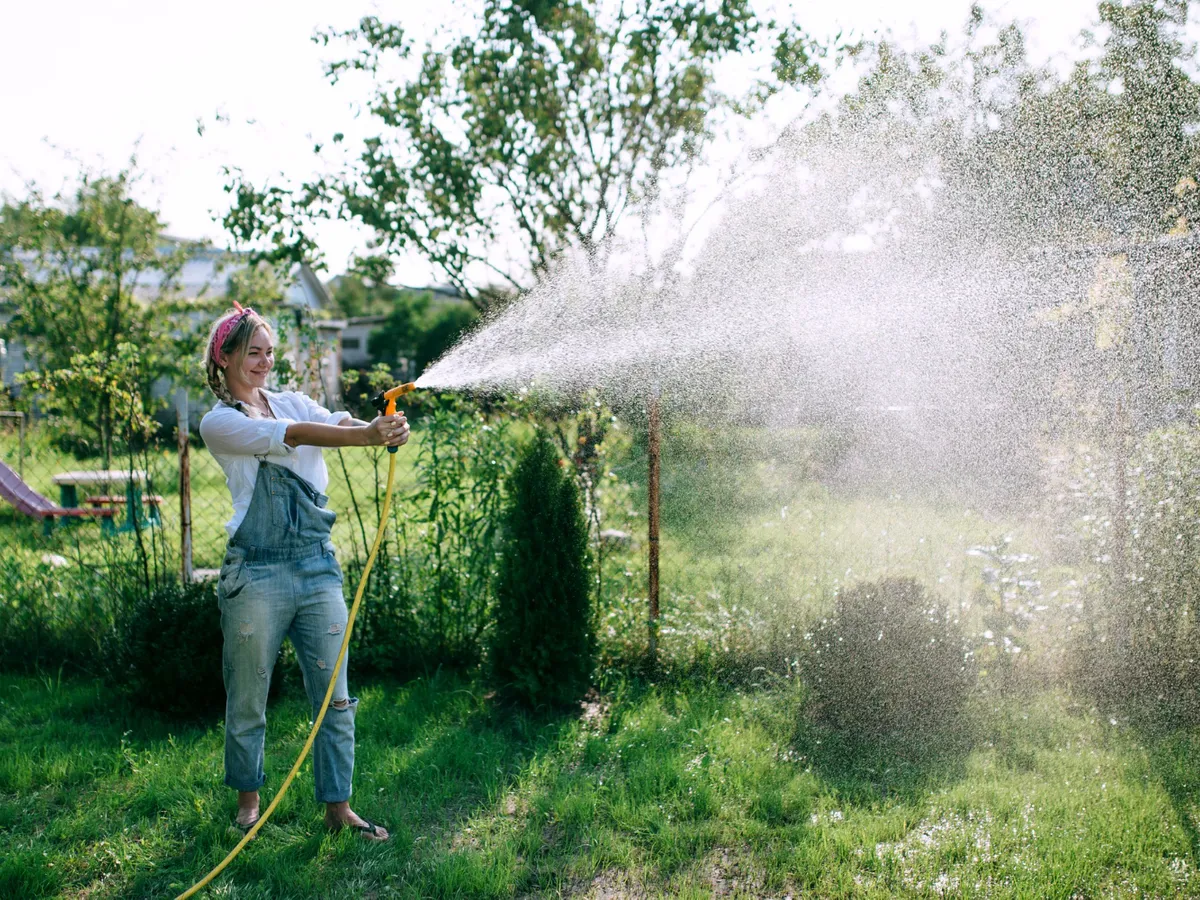 O tânără femeie într-o cămașă albă și o salopetă din denim udă gazonul - Foto: Magnific.com (Imagine cu rol ilustrativ)
