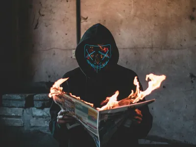 Persoană purtând o mască neagră citeşte dintr-un ziar care arde - Foto: Pexels/Connor Danylenko