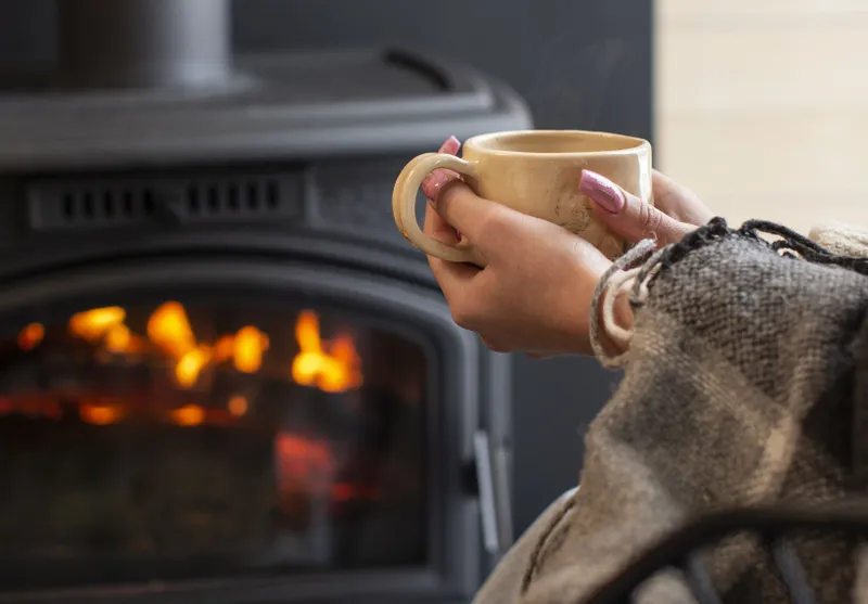 Femeie relax&acirc;ndu-se &icirc;n faţa şemineului, cu o cană de cafea fierbinte - Foto: Magnific.com/magnific