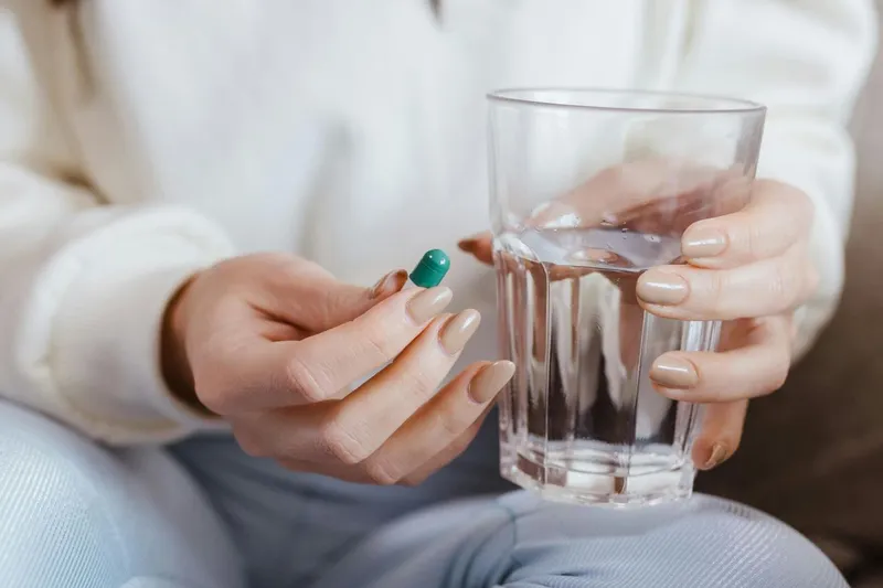 Tânără femeie pregătindu-se să bea cu apă, dintr-un pahar, o pastilă, pe care o ţine între degete - Foto: Pexels/Jonathan Borba